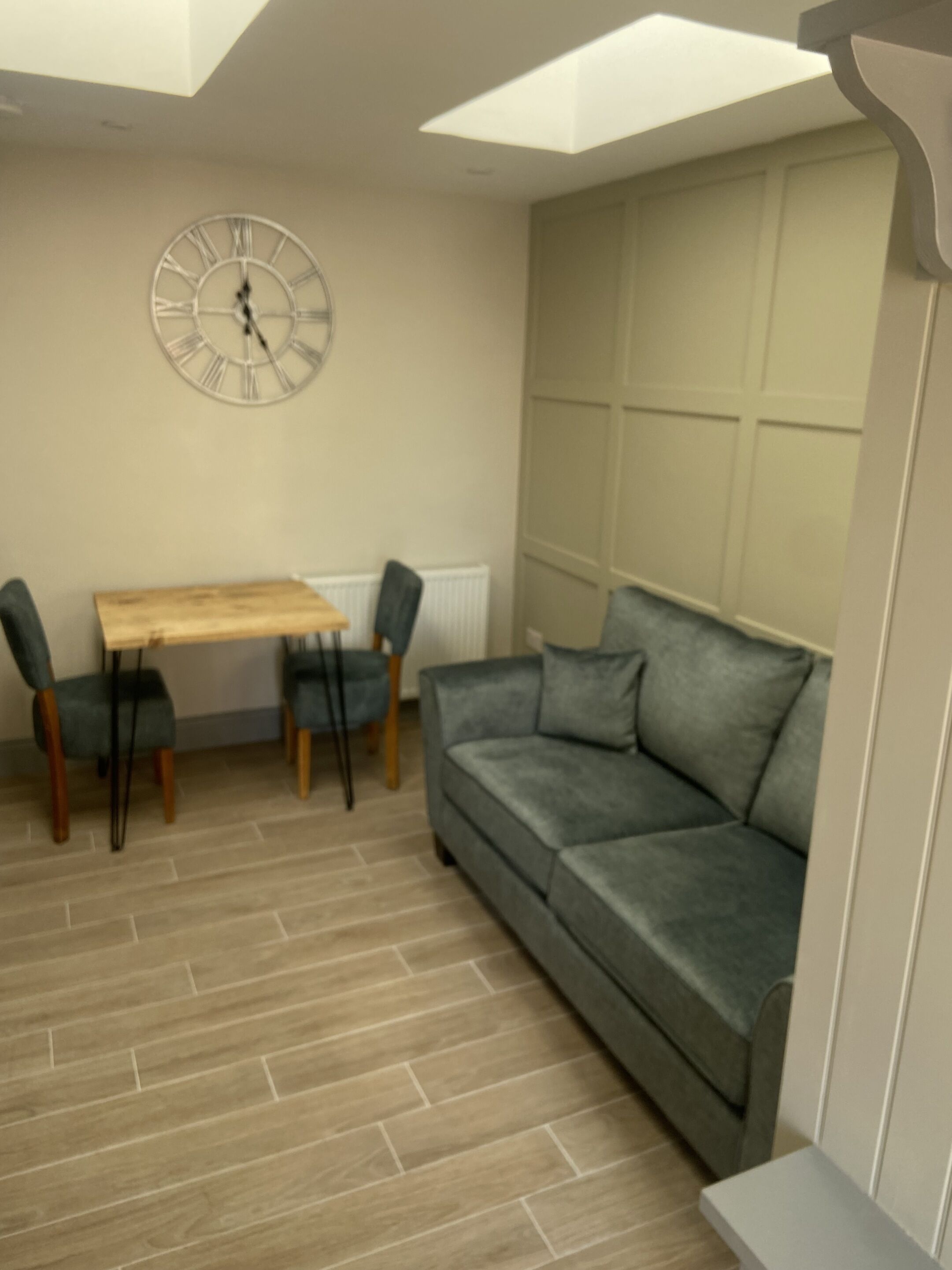 A room with a grey sofa, a wooden dining table with two chairs, and a wall clock.