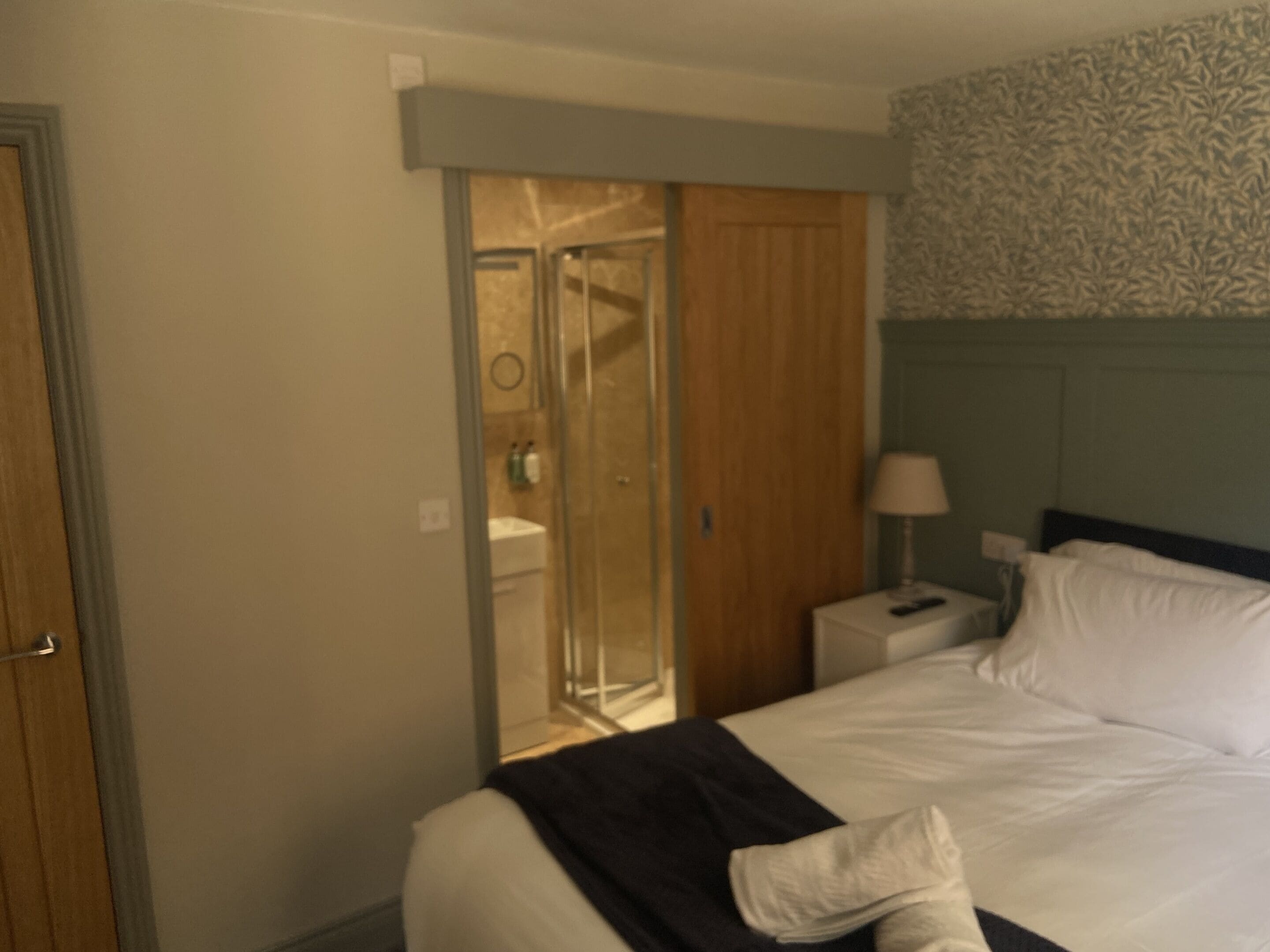 Habitación de hotel con cama, mesita de noche, lámpara, baño con ducha y puerta que da a otra habitación.