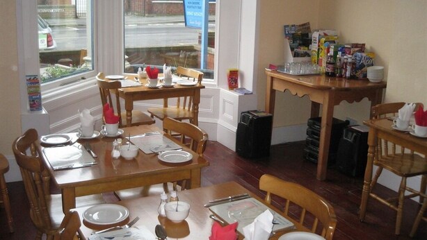 Un espace repas avec des tables et des chaises en bois, une petite table avec des condiments, et une fenêtre sur laquelle est affiché un panneau indiquant « COMPLET ».