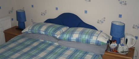 A bedroom with a bed, two pillows, a nightstand with a lamp, a coffee maker, and a small table with toiletries.