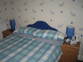 Un dormitorio con cama, dos almohadas, una mesita de noche con lámpara, cafetera y una pequeña mesa con artículos de aseo.