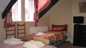 Une chambre avec deux lits, une chaise en bois, un radiateur, une commode noire et une fenêtre avec des rideaux.