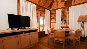 A wooden-floored room with a TV on a wooden cabinet, wicker chairs around a table, and large windows with curtains.