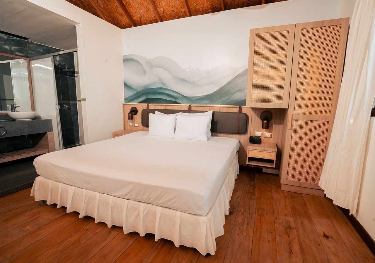 A bedroom with a large bed, wooden headboard, a nightstand, a wall-mounted lamp, and a large window with a view of the outdoors.