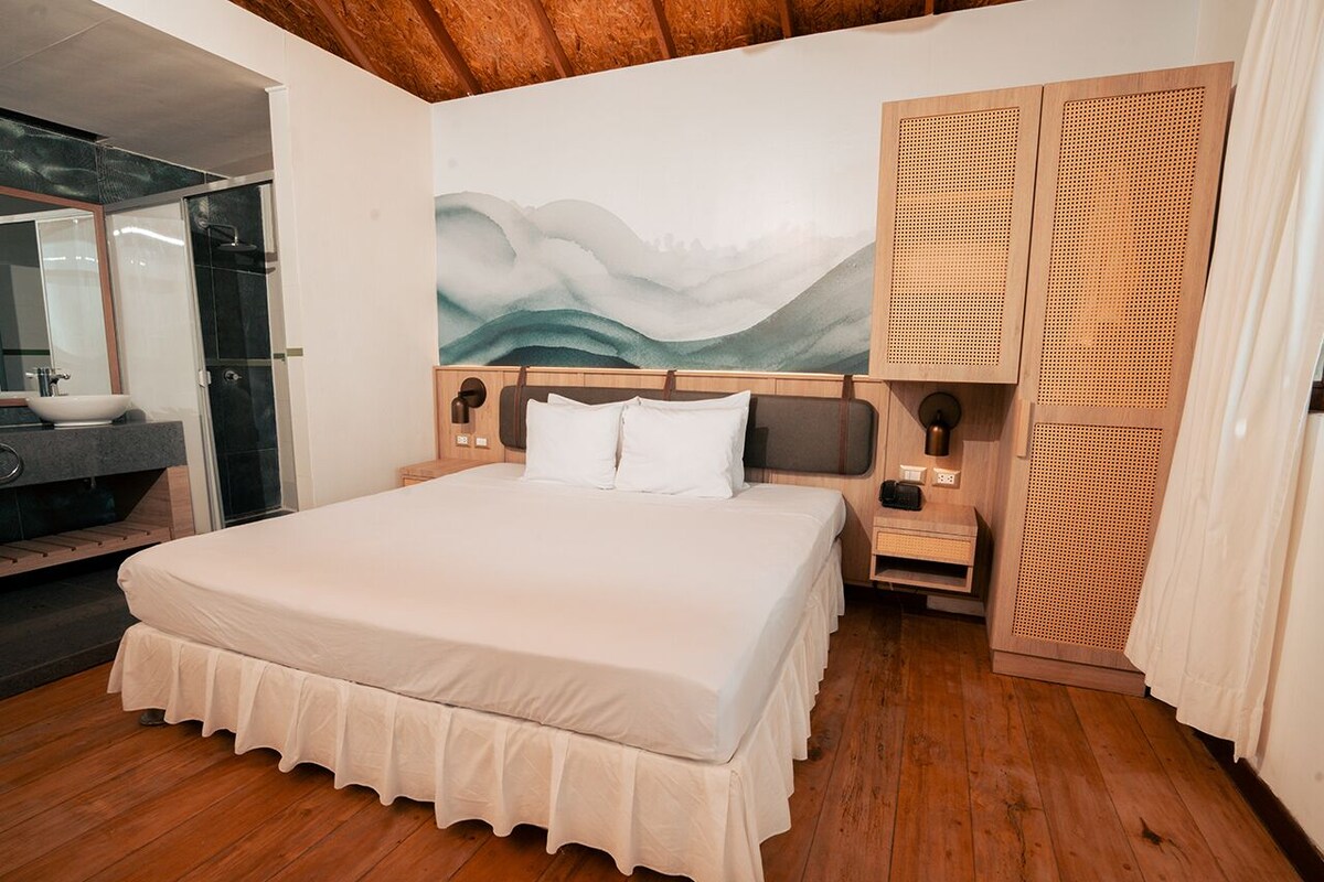 A bedroom with a large bed, wooden headboard, a nightstand, a wall-mounted lamp, and a large window with a view of the outdoors.