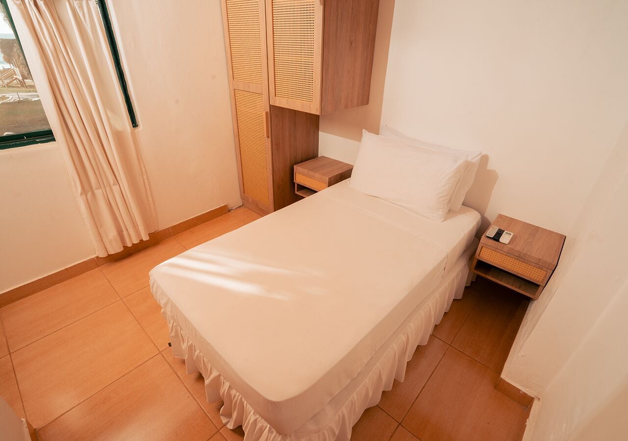 A neatly made bed in a room with a wooden nightstand, a small wooden cabinet, and a window with curtains.