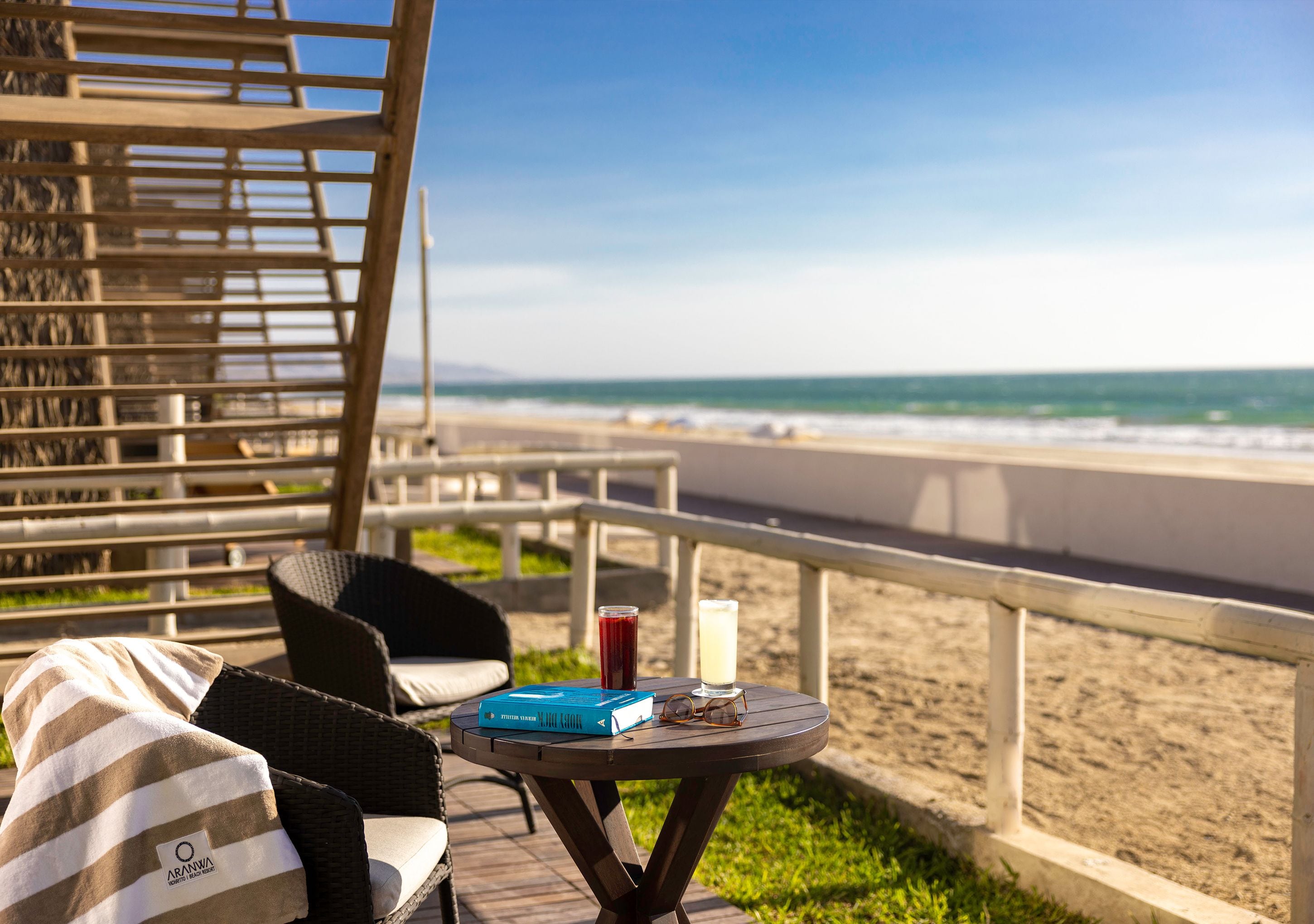 Un espacio de descanso frente al mar, con vistas al océano, una mesa con una bebida y un libro, y una silla de mimbre con una toalla a rayas.