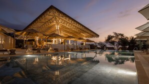Une piscine entourée de chaises longues et de parasols, elle-même bordée d’un bâtiment doté d’un toit de chaume.