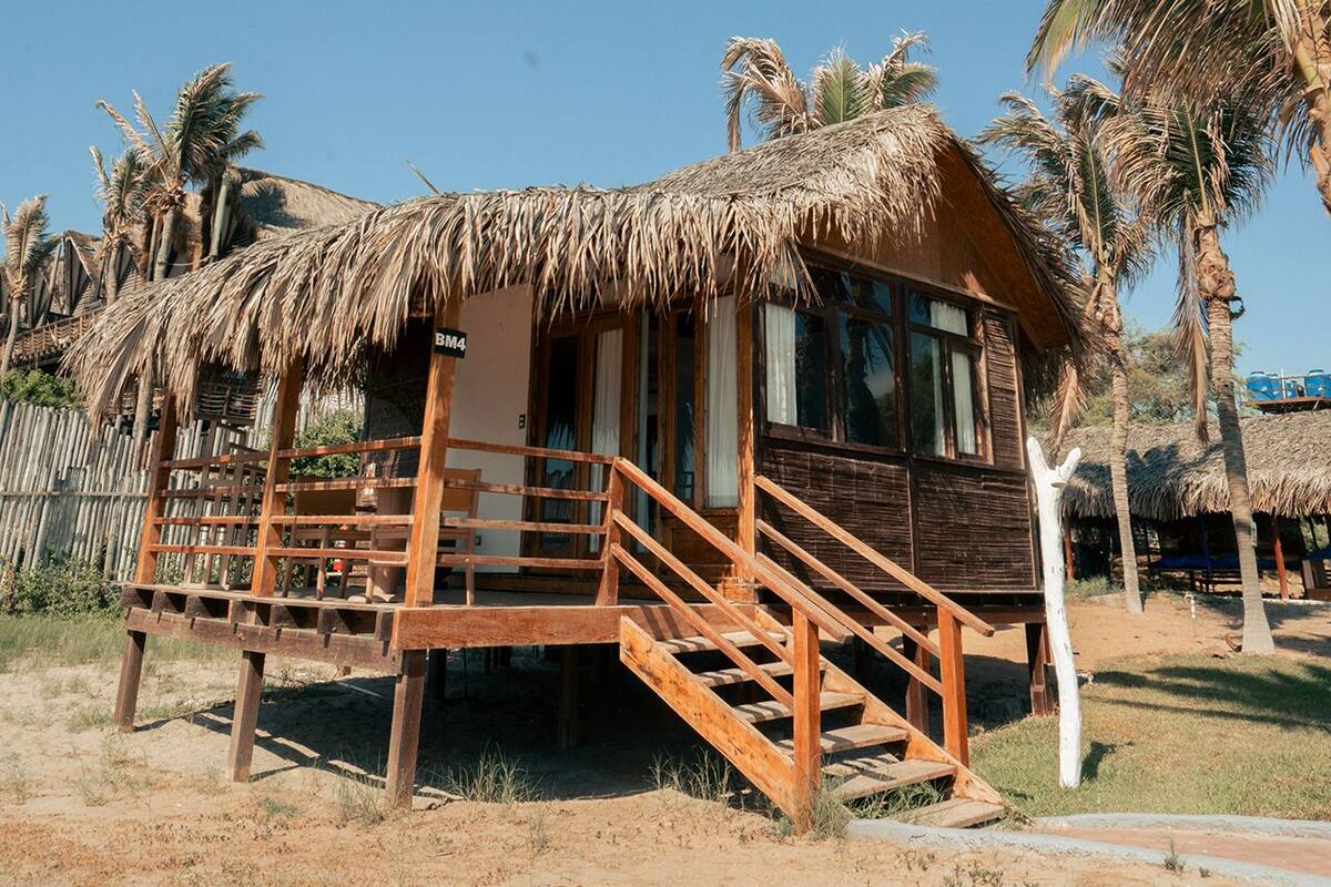 Una cabaña con techo de paja y una escalera de madera que lleva a la entrada, rodeada de palmeras.