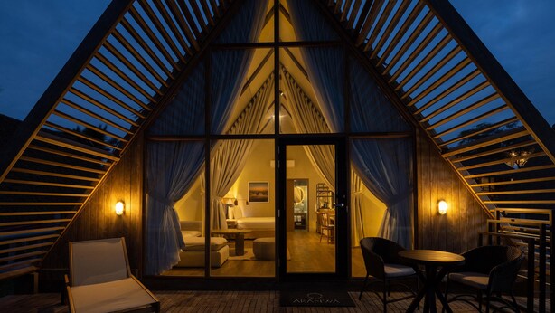 A triangular wooden structure with a glass entrance, a bed, a table, and chairs visible inside.