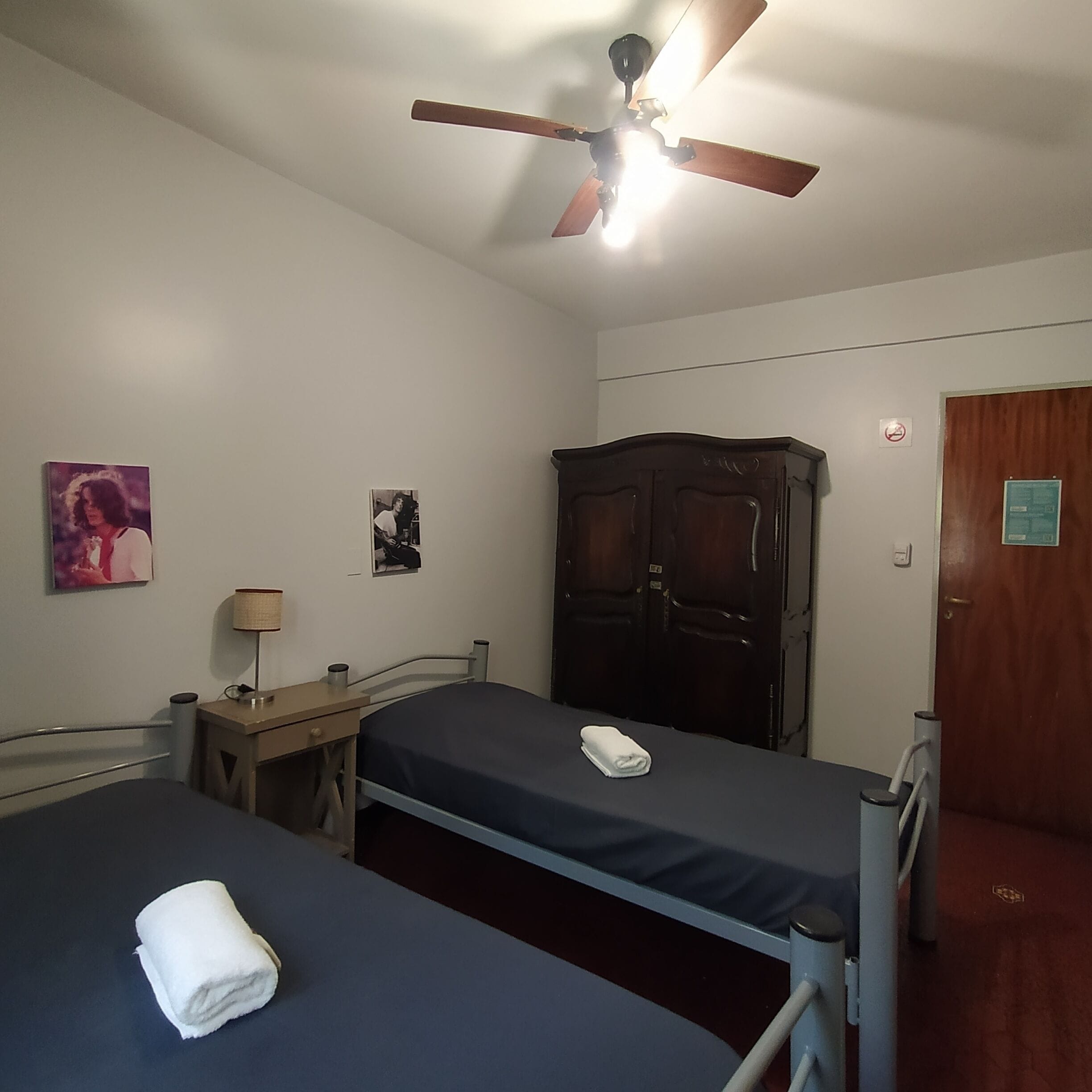 A room with two single beds, a desk, a chair, a wardrobe, a ceiling fan, and a wall-mounted lamp.