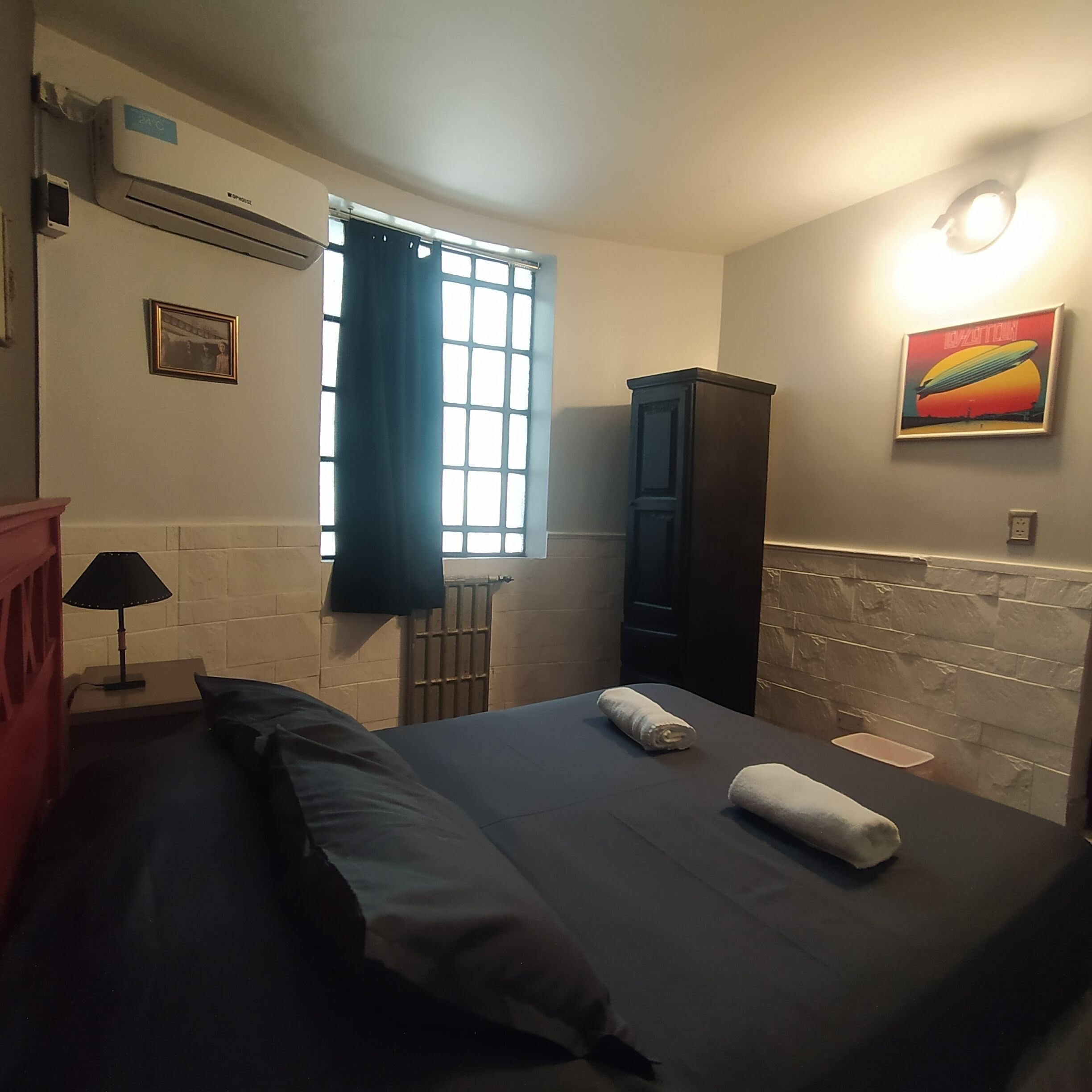 A bedroom with a bed, a nightstand, a lamp, a window with curtains, a radiator, a cabinet, and a wall-mounted light fixture.