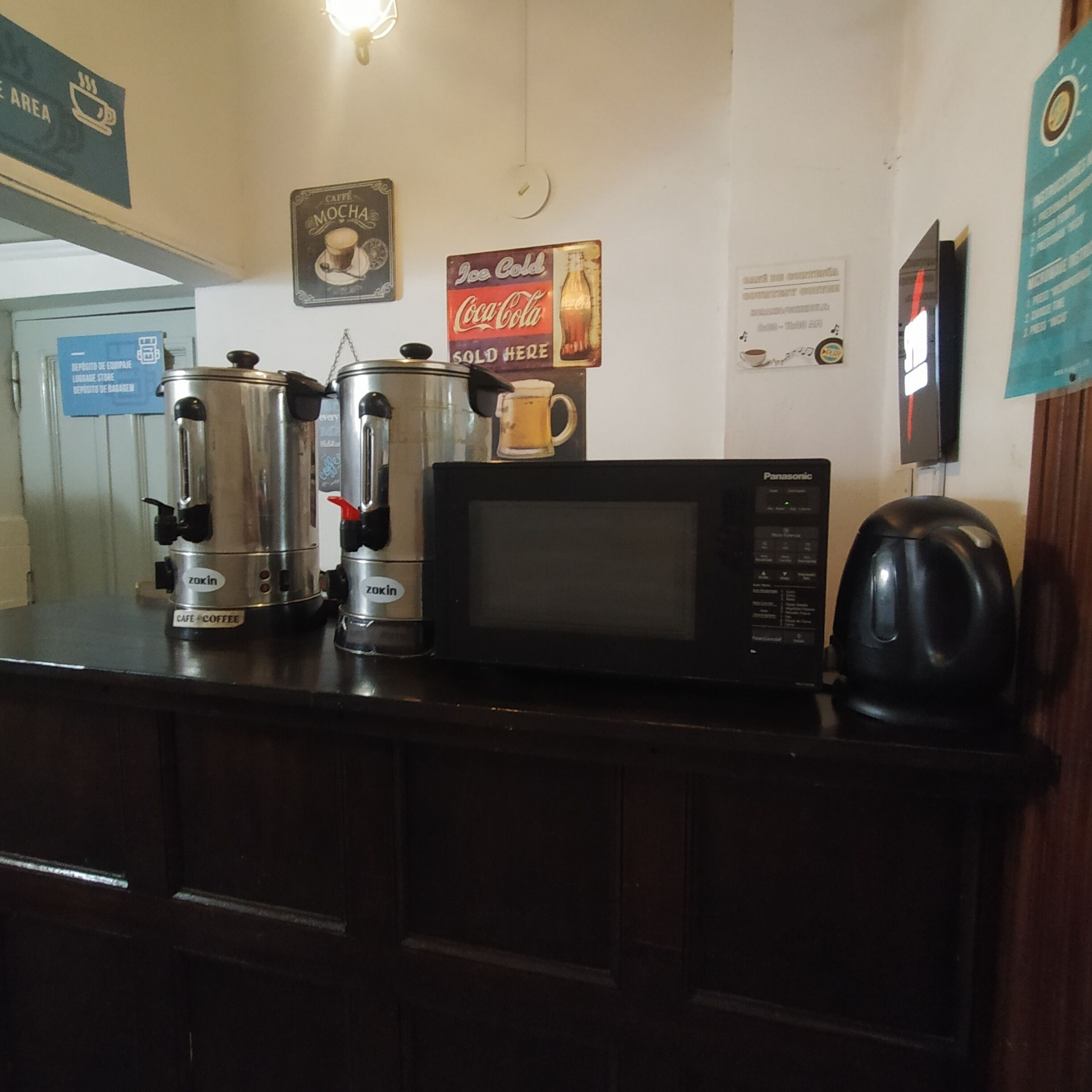 A kitchen counter with a coffee maker, a microwave, and a kettle. There are signs on the wall indicating a coffee area and a Coca-Cola sign.