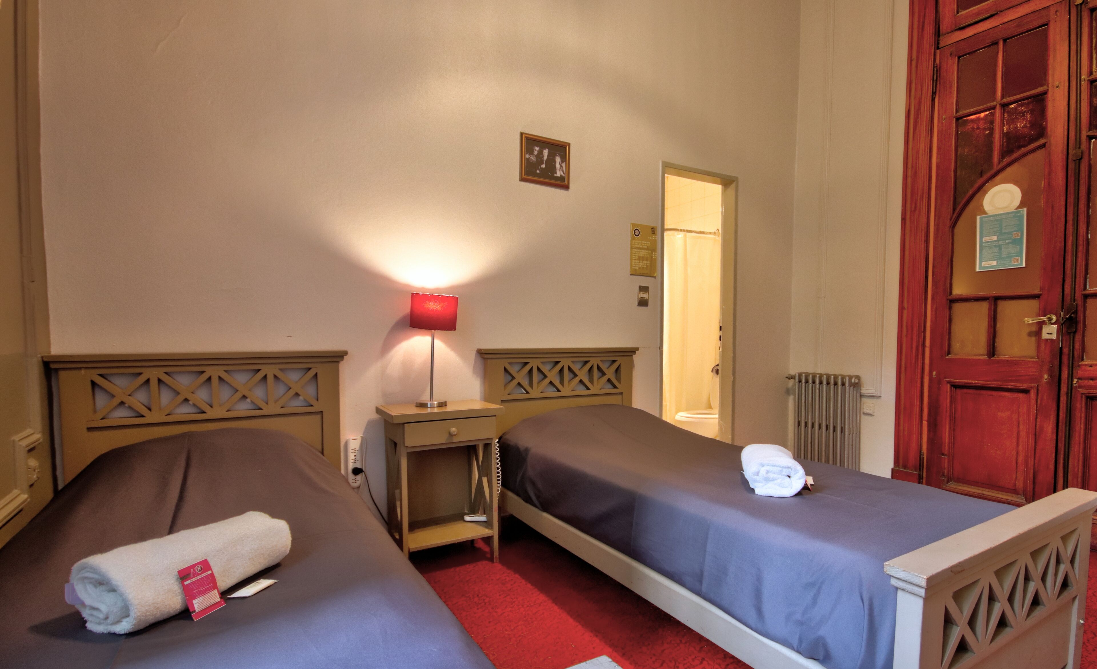 Quarto de hotel com duas camas de solteiro, mesa de cabeceira com abajur, tapete vermelho, porta de madeira e pequena moldura com quadro na parede.