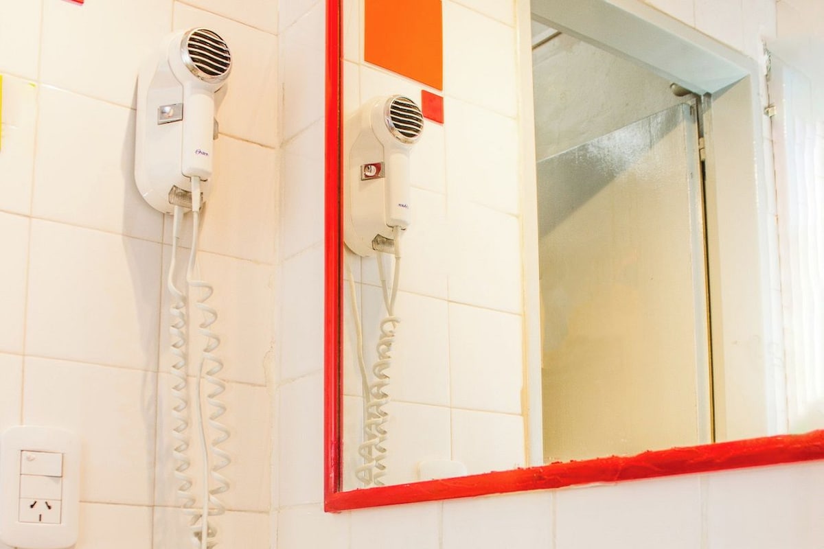 Un baño con un lavabo blanco, dos grifos, un espejo con marco rojo y dos secadores de pelo fijados en la pared.