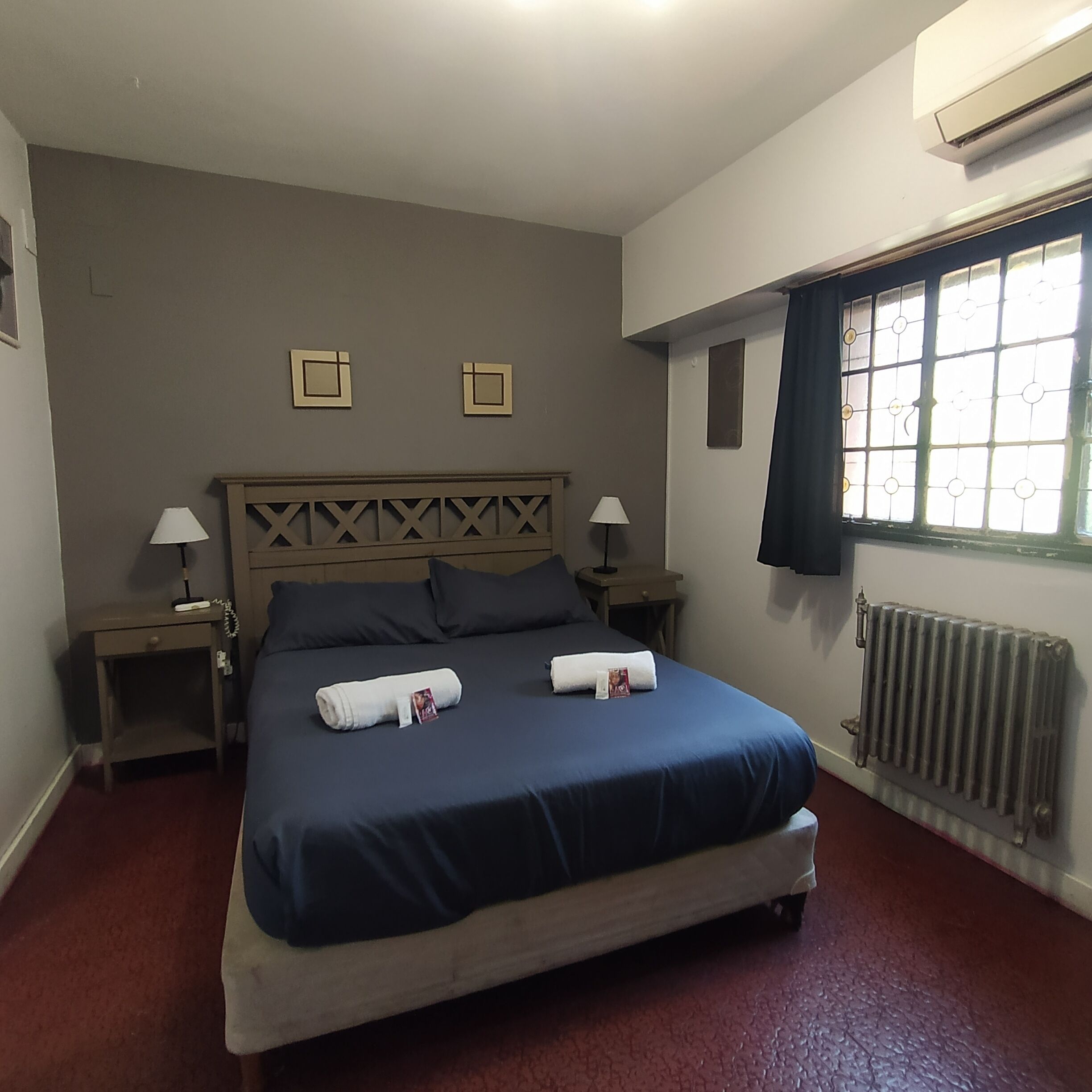 A hotel room with a large bed, two bedside tables with lamps, a radiator, and a window with curtains.