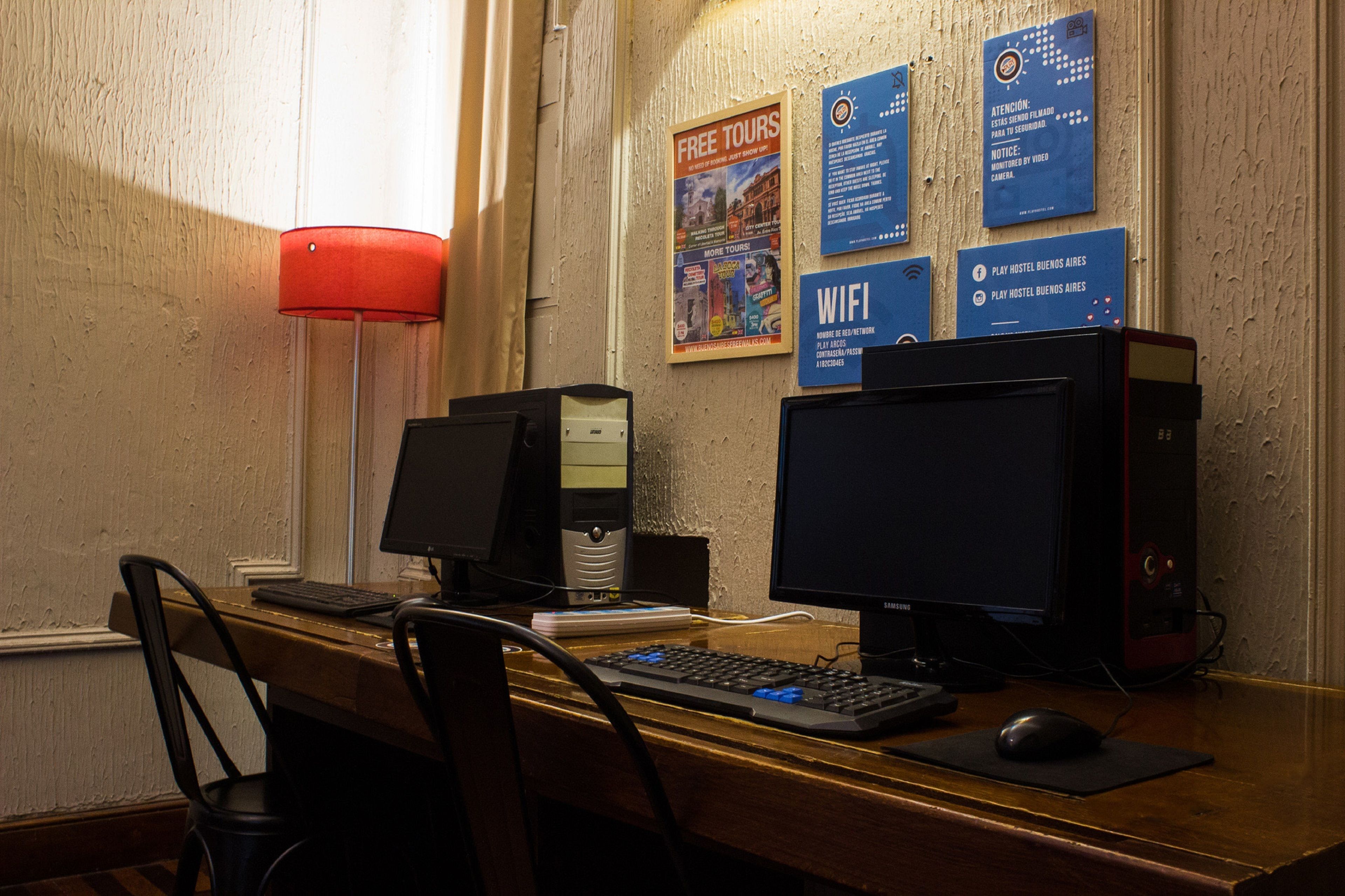 Een computerbureau met twee schermen, een toetsenbord en een muis. Op de muur hangen posters die gratis rondleidingen en WiFi aanprijzen.