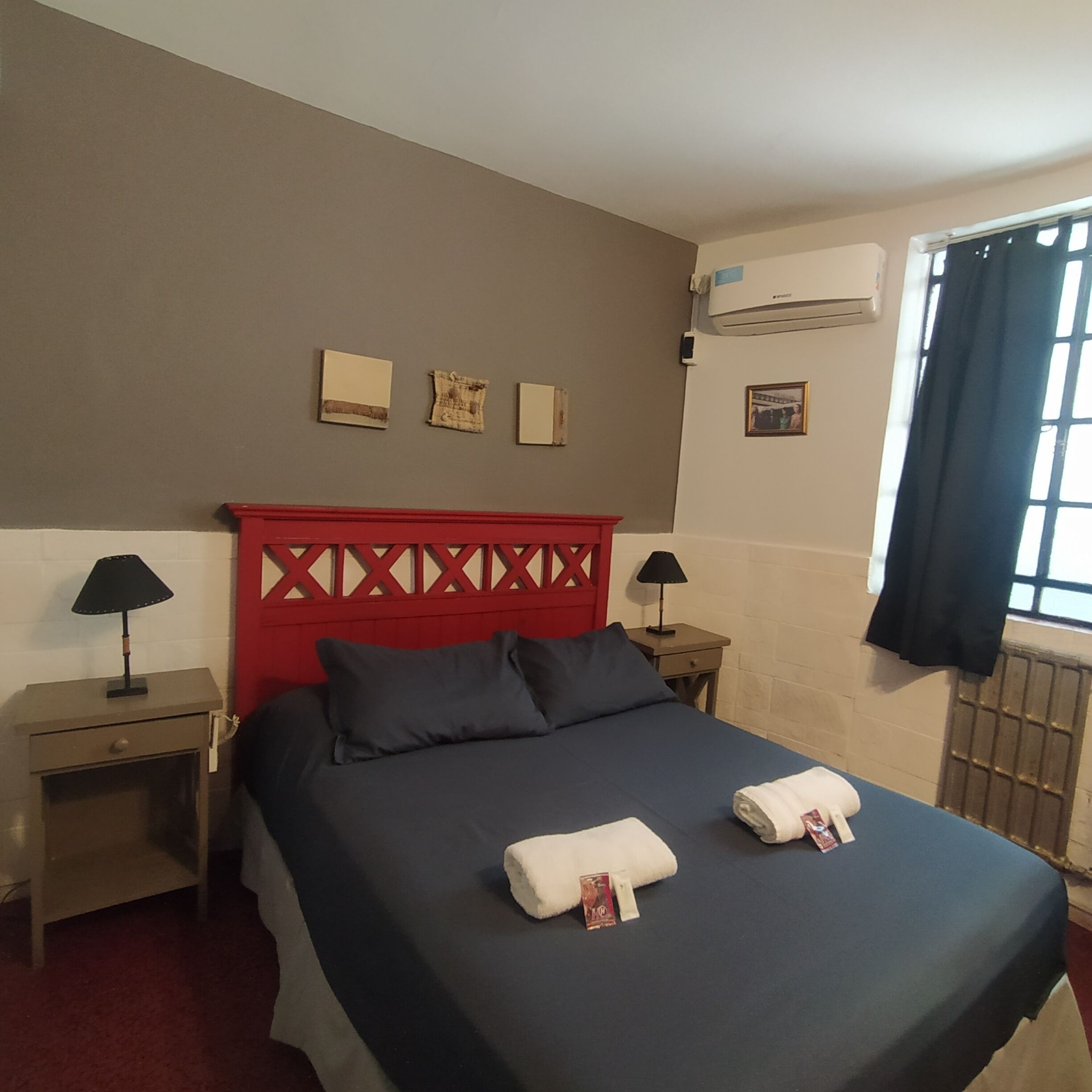 Una habitación de hotel con cama, dos mesitas de noche, cabecera roja y ventana con cortinas.