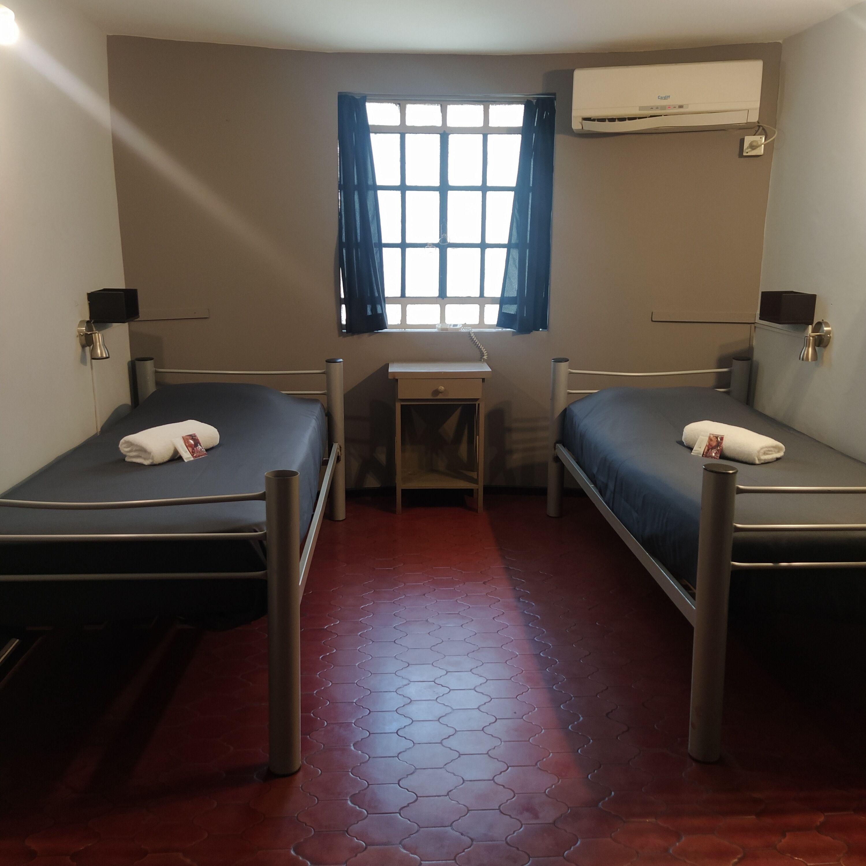 Una habitación con dos camas, un escritorio, un ventanal con cortinas, aire acondicionado y suelo de baldosas rojas.