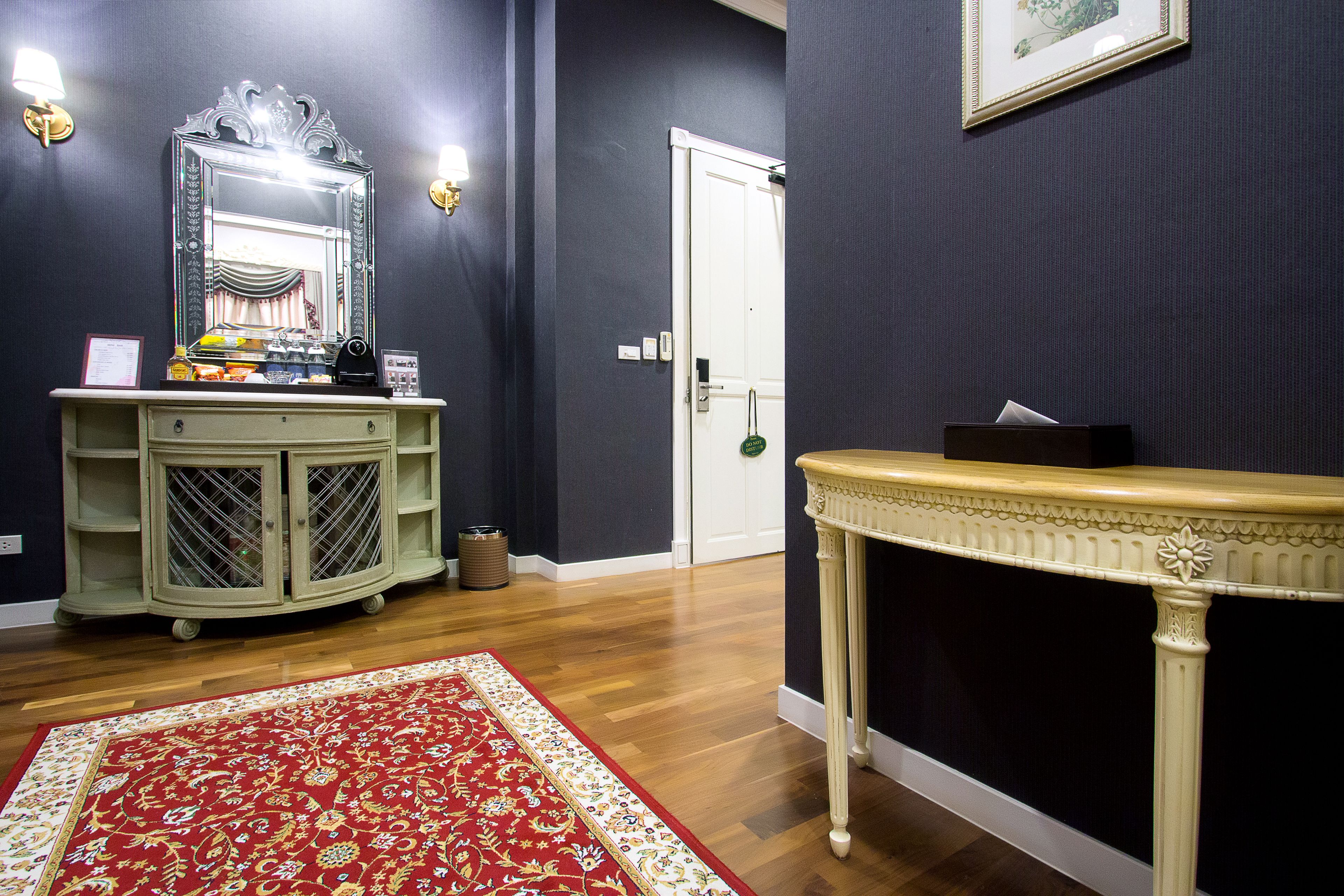Een kamer met een donkere wand, een houten vloer, een crème kleurige console tafel, een spiegel met decoratieve lijst, een rood tapijt met patroon, en een witte deur.