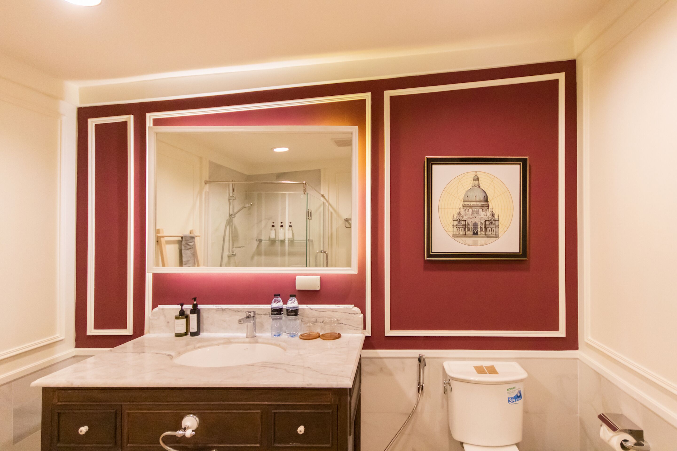 A bathroom with a marble sink, a large mirror, a framed picture of a building on the wall, and a toilet with a paper towel dispenser.