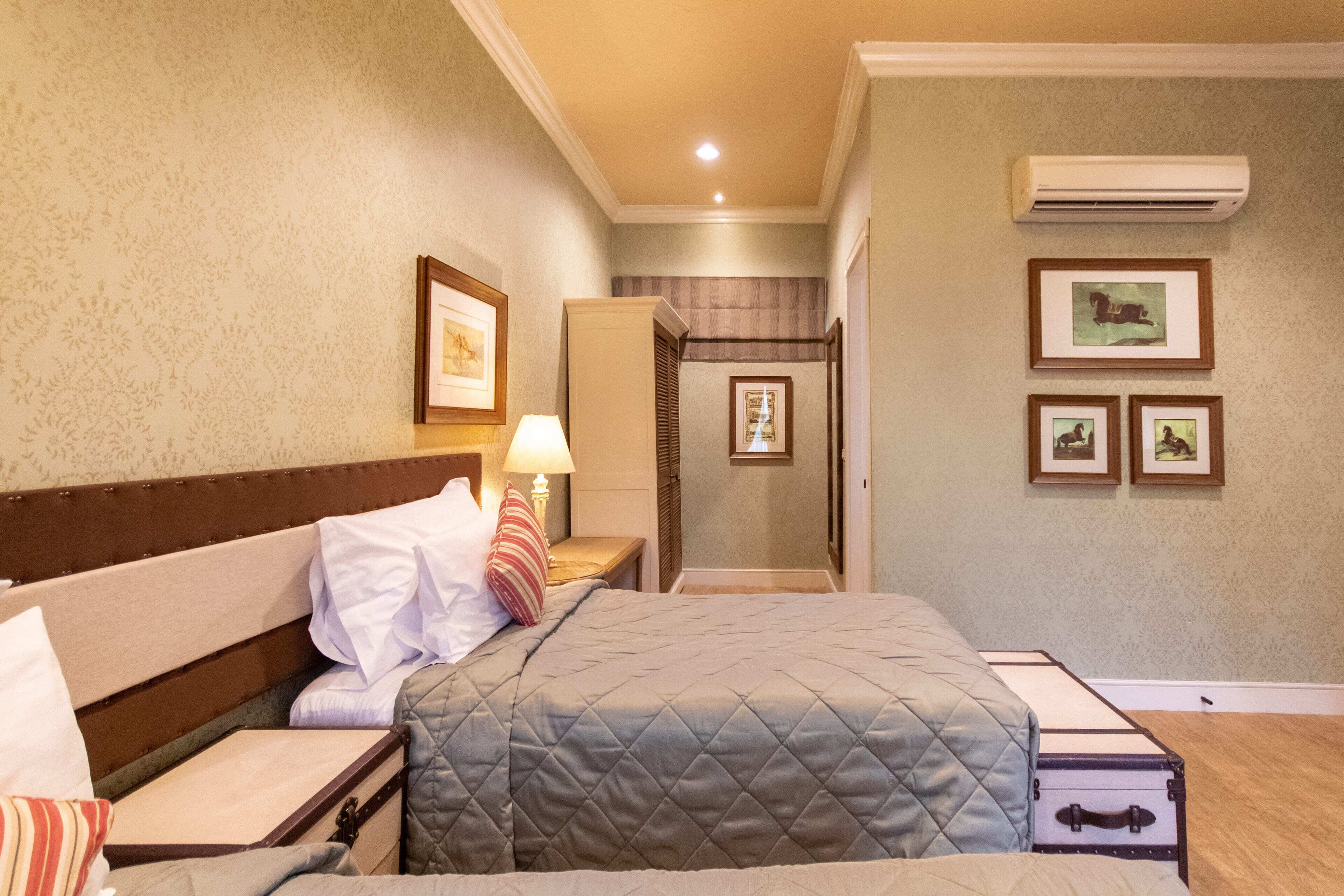 A hotel room with a bed, bedside tables, a lamp, framed pictures on the wall, and an air conditioning unit.