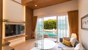 A modern living room with a flat-screen TV, a glass coffee table, and a sofa with cushions. There is a large window with curtains, a view of a swimming pool, and a wooden ceiling. - Rancho Charnvee Khaoyai (Pak Chong)