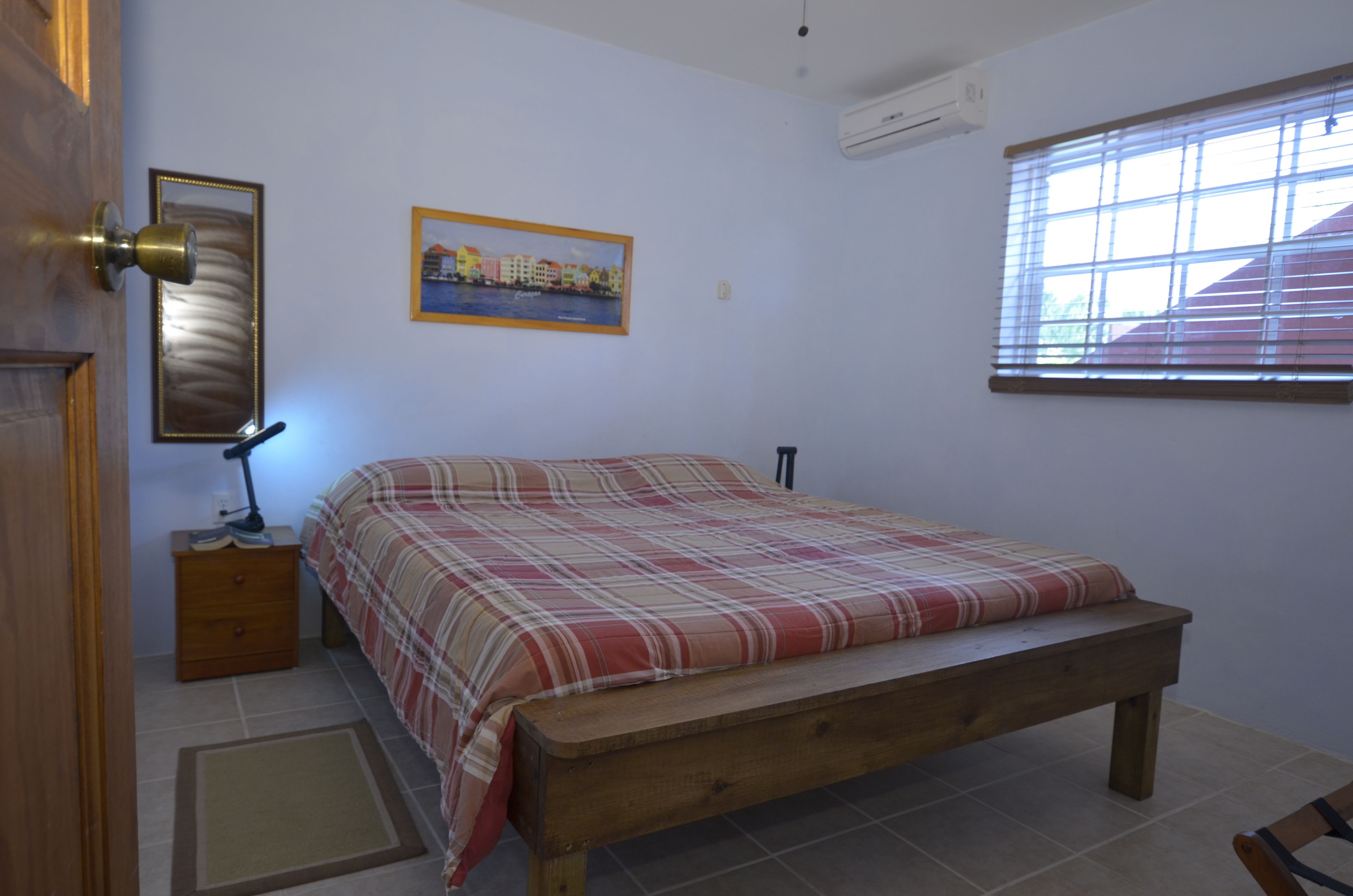 A bedroom with a wooden bed, a nightstand, a lamp, a framed picture on the wall, and a window with blinds.