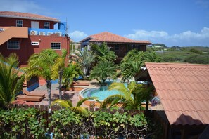 A resort with a swimming pool, palm trees, and a red-roofed building. - Nos Krusero Apartments (Sabana Westpunt)