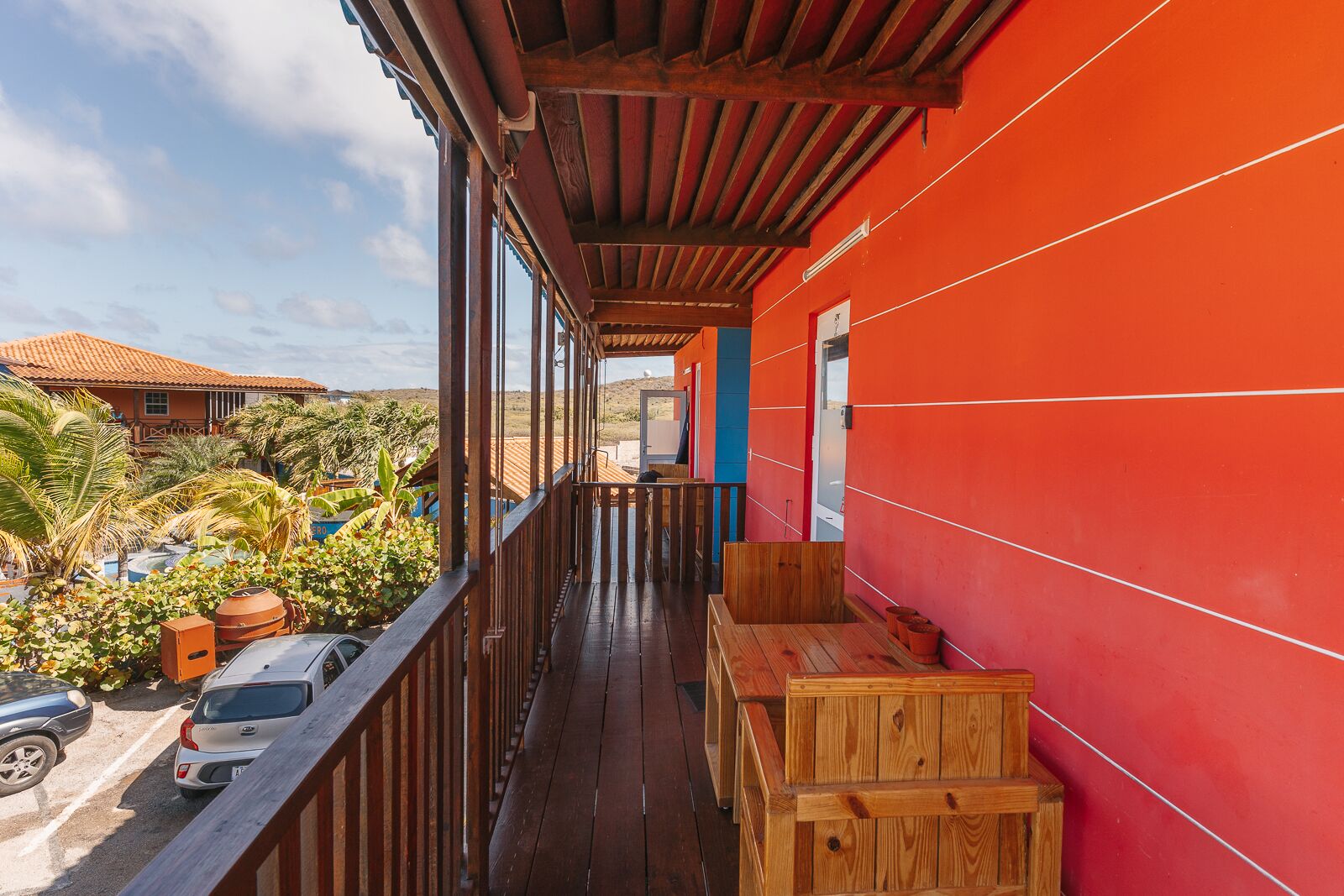 Un balcon avec des balustrades en bois, un mur rouge et une vue sur des palmiers et un bâtiment en arrière-plan.