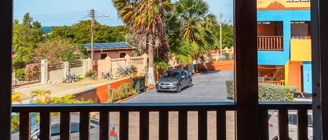 A view from a balcony with a wooden railing, overlooking a street with parked cars and palm trees.