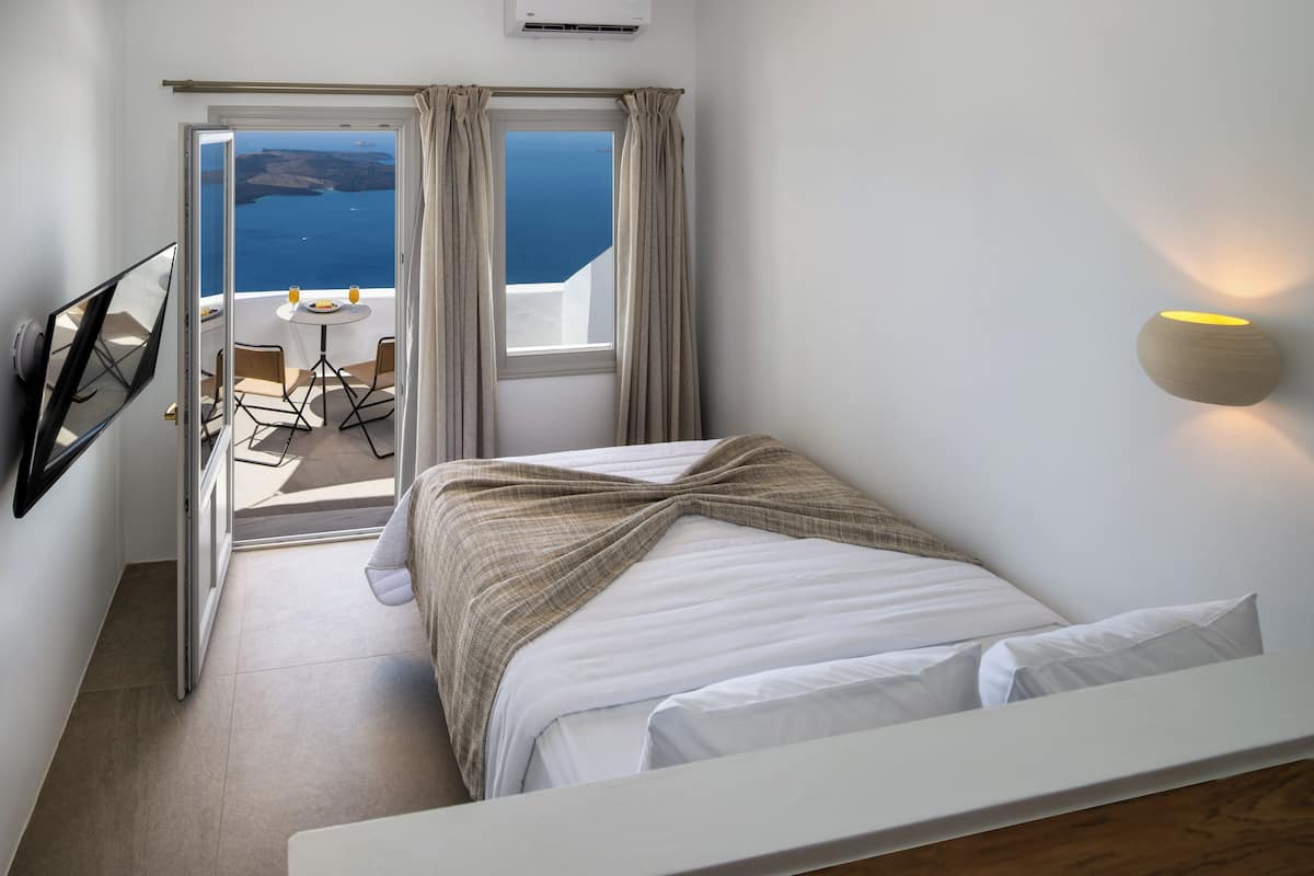 A bedroom with a bed, a small table with chairs, a view of the sea, and a wall-mounted light fixture.