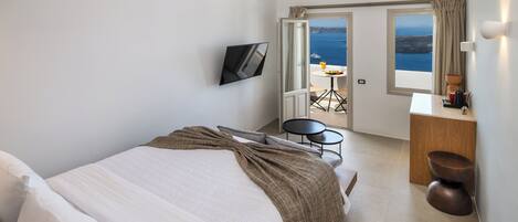 A hotel room with a bed, a TV, a desk with a chair, and a view of the sea.