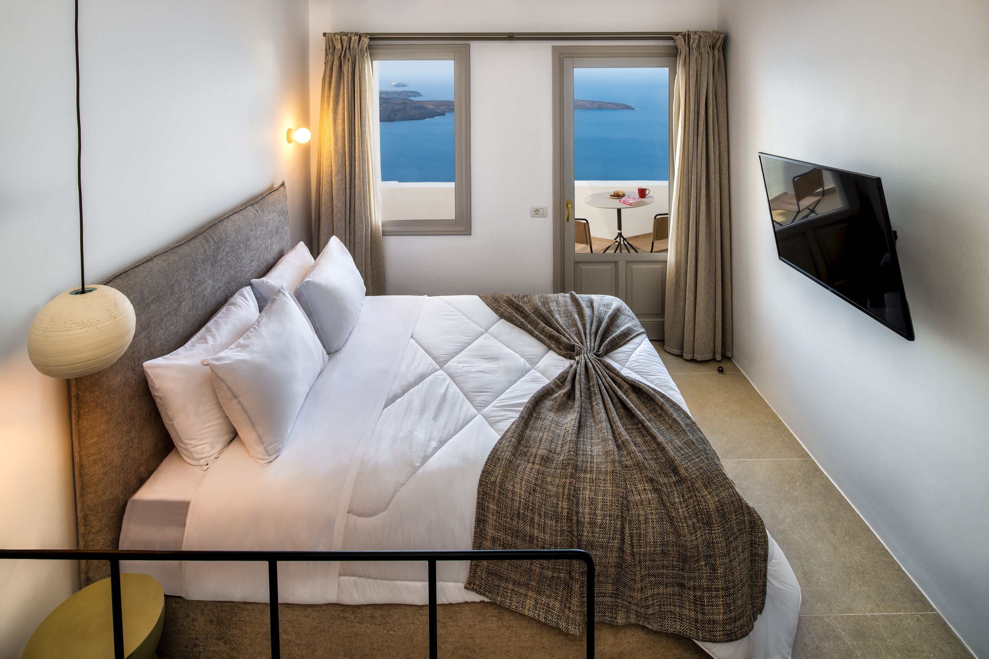 Ein ordentlich bezogenes Bett mit Meerblick durch ein Fenster, ein Flachbildfernseher an der Wand und ein runder Tisch mit Stühlen draußen.