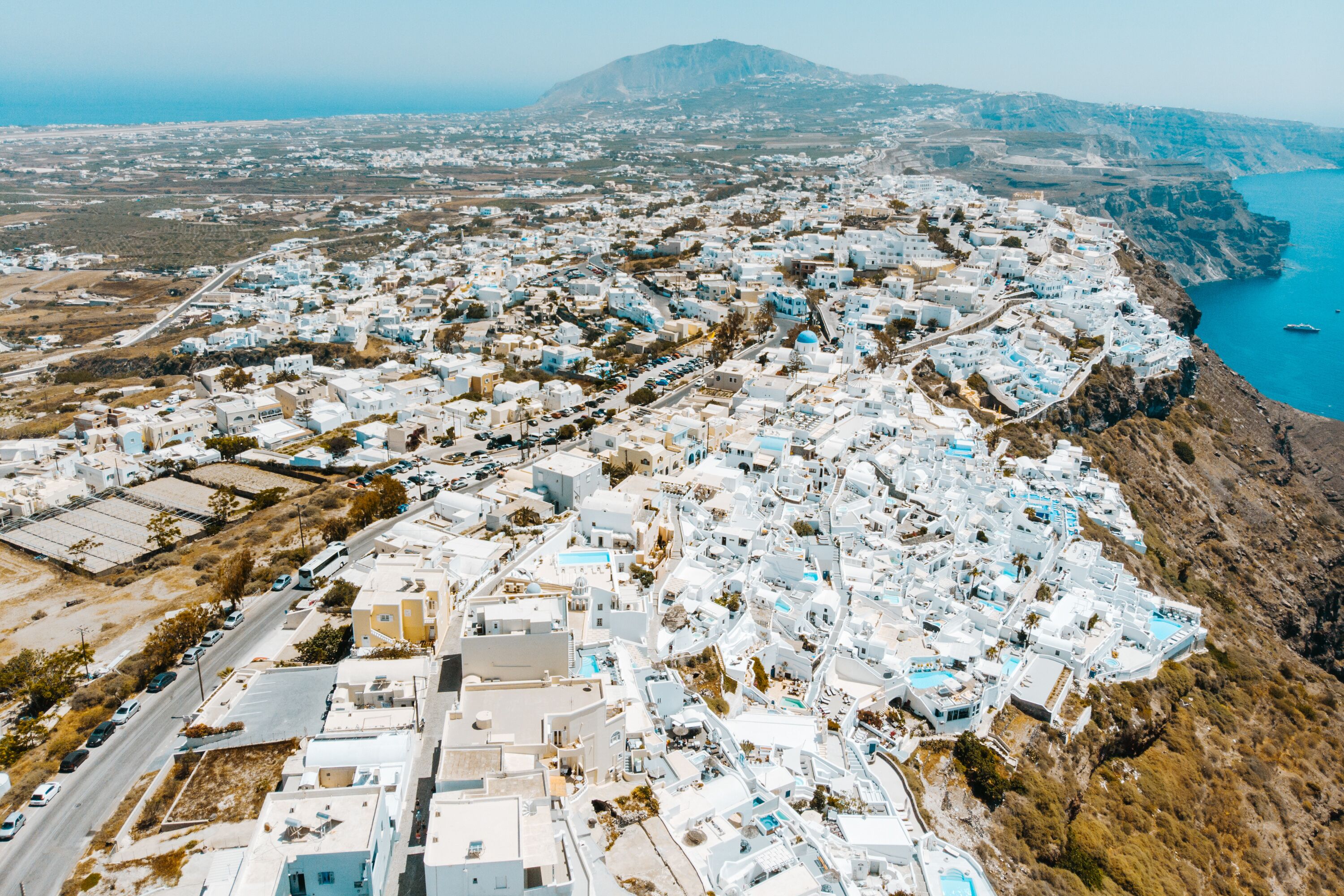 An aerial view of a coastal town with white buildings, a clear blue sea, and a mountainous backdrop.