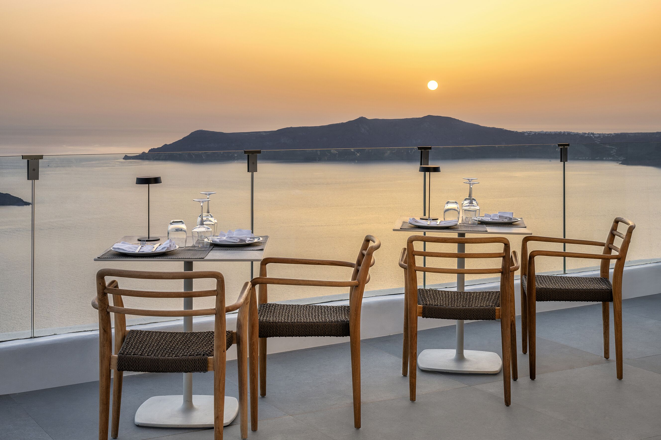 Una terraza con una mesa lista para dos personas, con vistas a un cuerpo de agua tranquilo y colinas lejanas durante el atardecer.