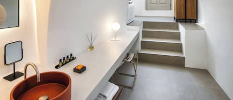 Un baño moderno con un lavamanos ovalado grande, un espejo y una lámpara. Hay toallas en un banco y un estante pequeño con artículos de tocador.