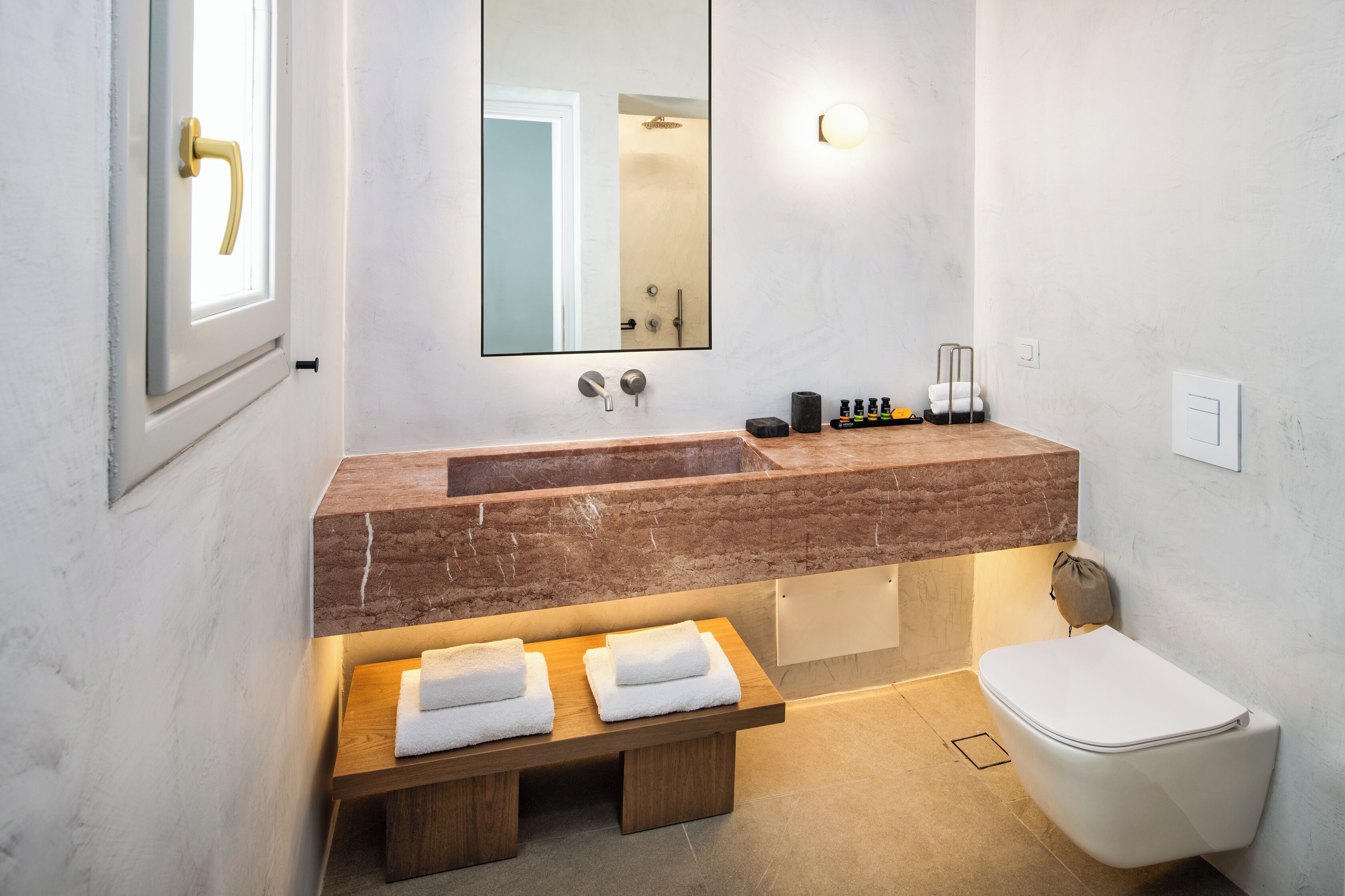 A modern bathroom with a marble sink, a wooden bench with towels, a white toilet, and a large mirror.