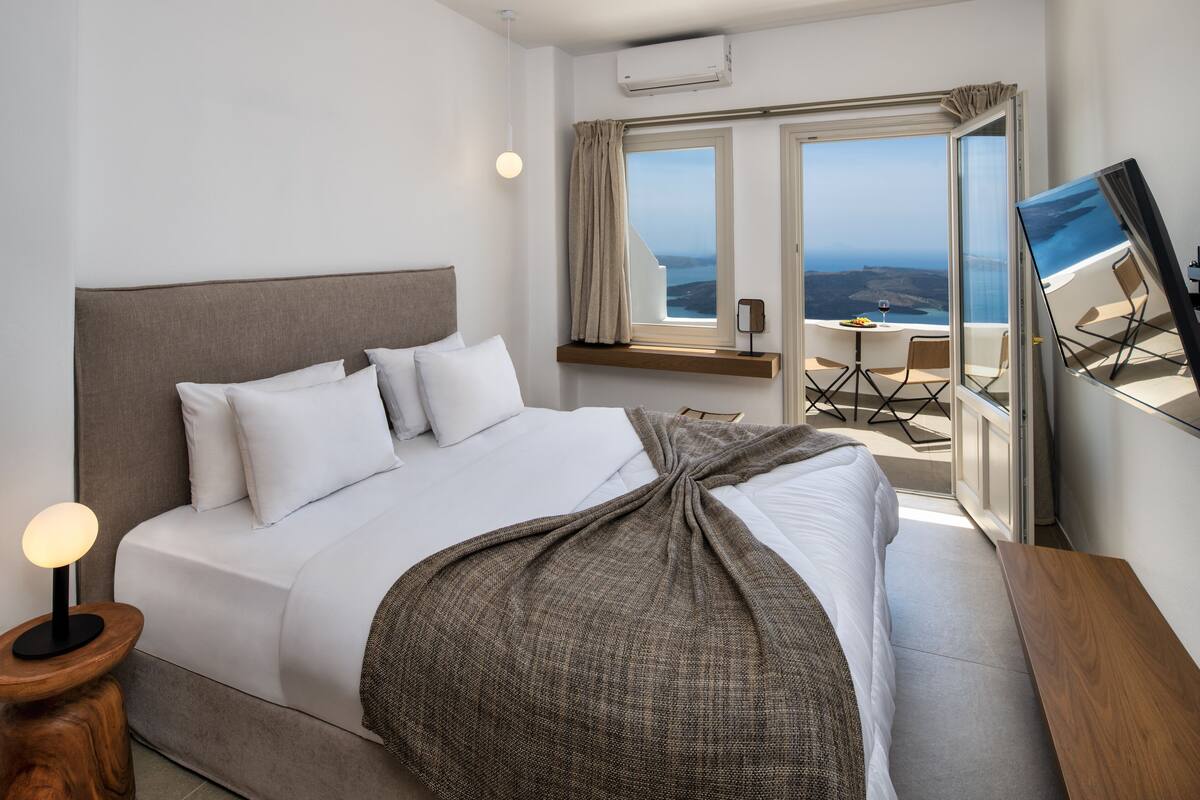 A modern hotel room with a large bed, a bedside table with a lamp, a window with a view of the sea, and a balcony with a table and chairs.