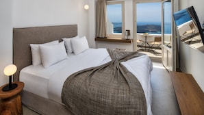 A modern hotel room with a large bed, a bedside table with a lamp, a window with a view of the sea, and a balcony with a table and chairs.