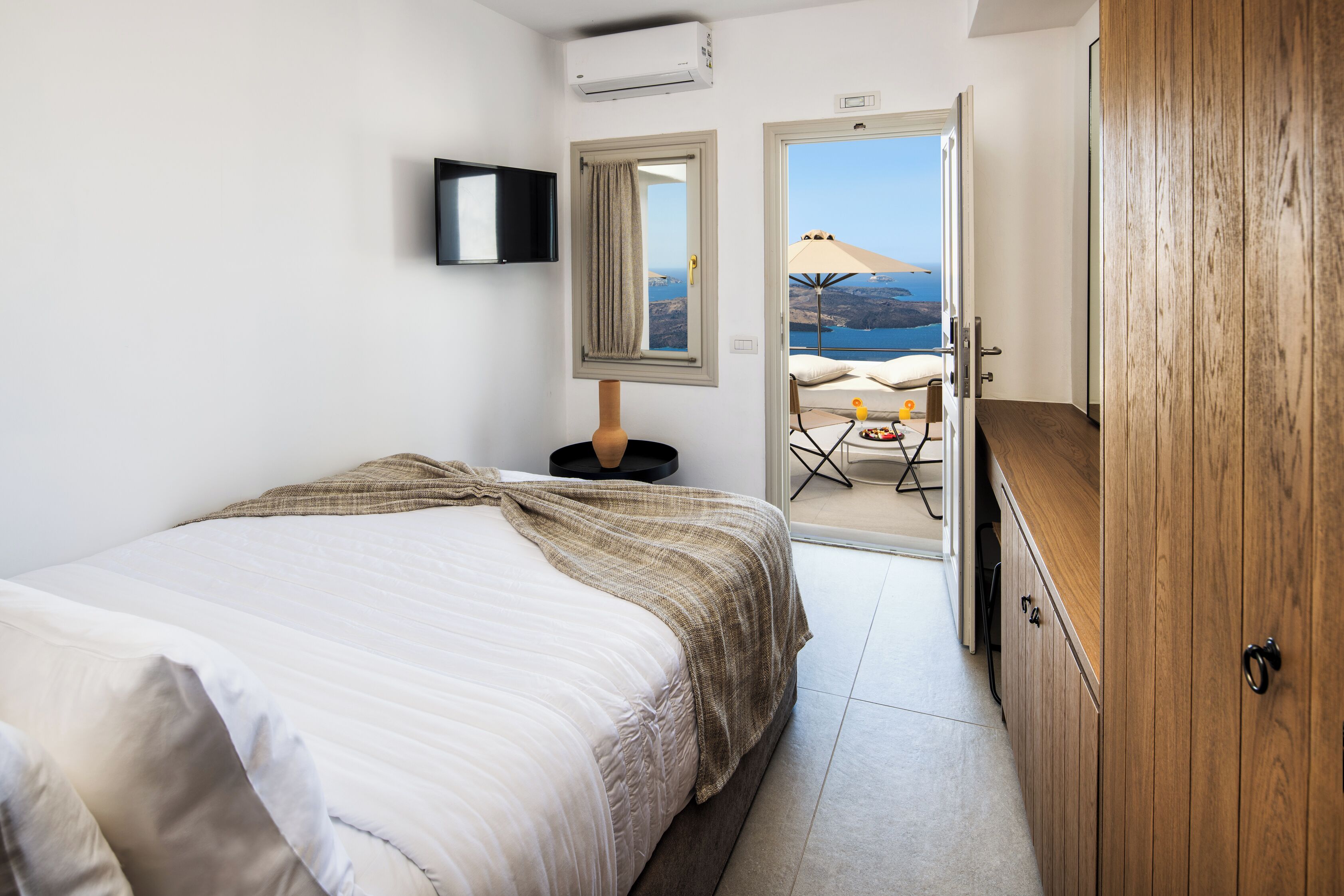 A bedroom with a bed, a wooden wardrobe, a TV mounted on the wall, and a view of the sea through an open door.