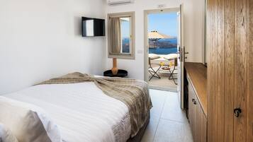 A bedroom with a bed, a wooden wardrobe, a TV mounted on the wall, and a view of the sea through an open door.