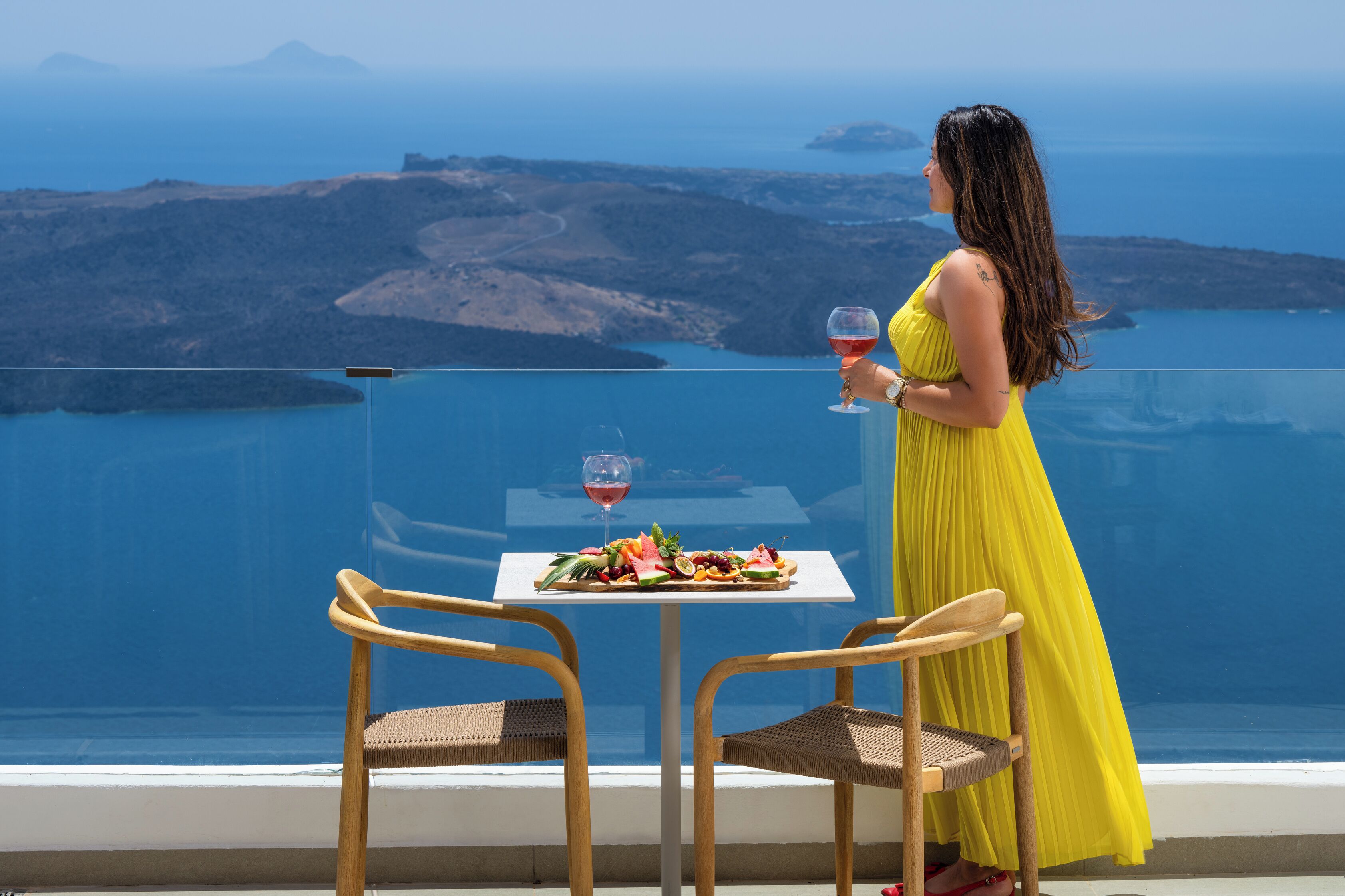Una mujer con un vestido amarillo sentada a una mesa con comida y una copa de vino tinto, con vistas a un paisaje costero.