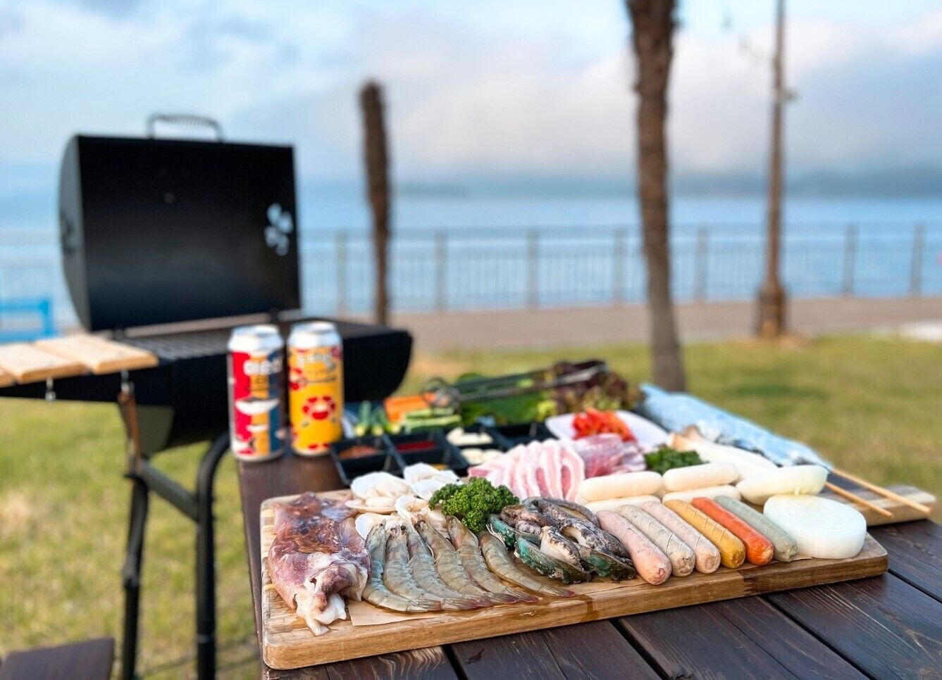 A picnic setup with a variety of meats and seafood on a wooden board, a grill, and a can of soda in the background.