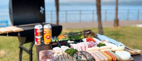 A picnic setup with a variety of meats and seafood on a wooden board, a grill, and a can of soda in the background.