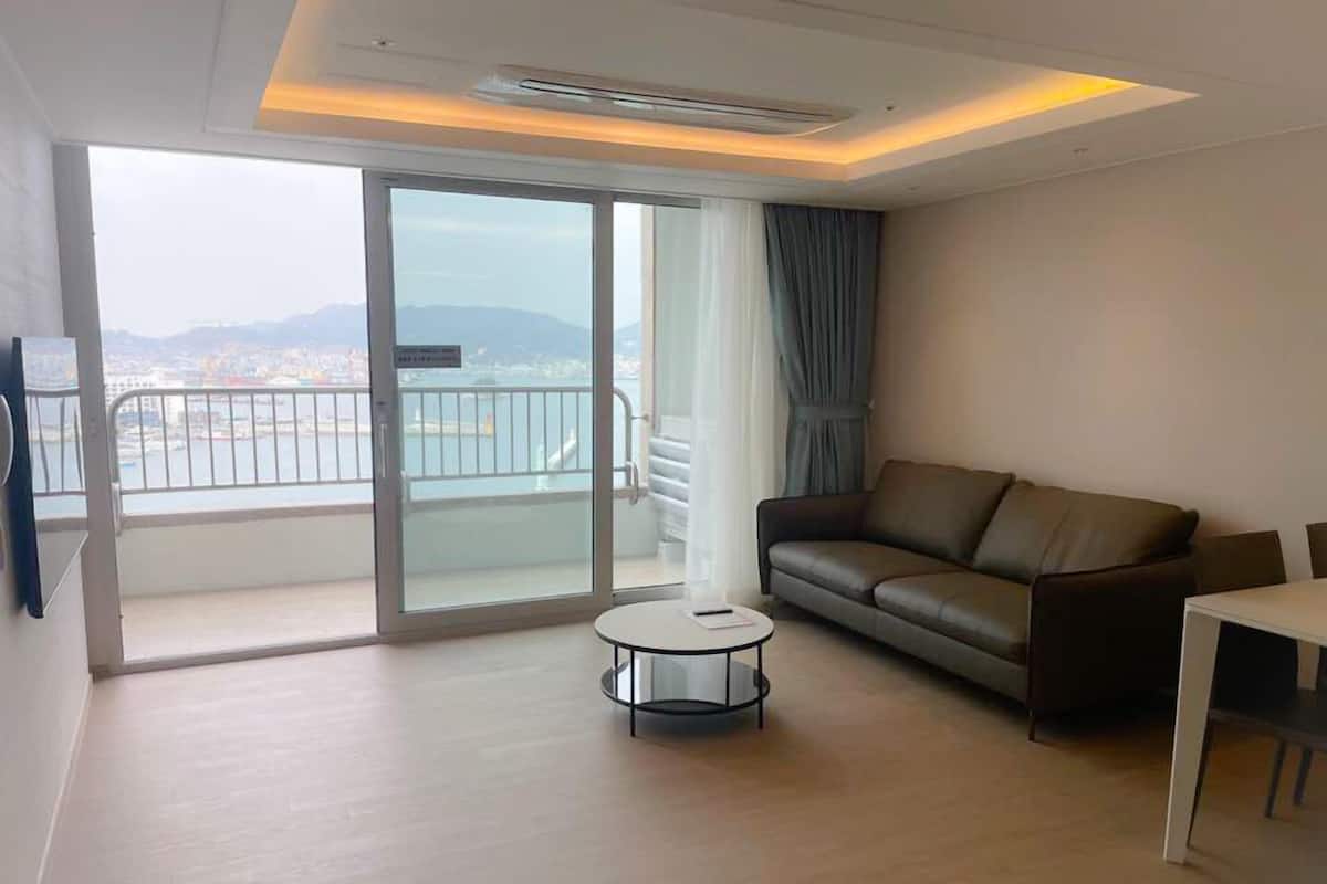 Un salon moderne avec un canapé en cuir, une petite table ronde et une grande porte-fenêtre coulissante donnant sur un balcon avec vue sur la ville.