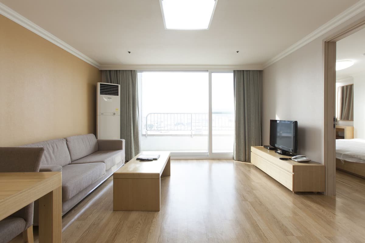 Une chambre d’hôtel moderne avec un canapé, une table basse en bois, un téléviseur posé sur un meuble en bois et une grande fenêtre avec des rideaux.