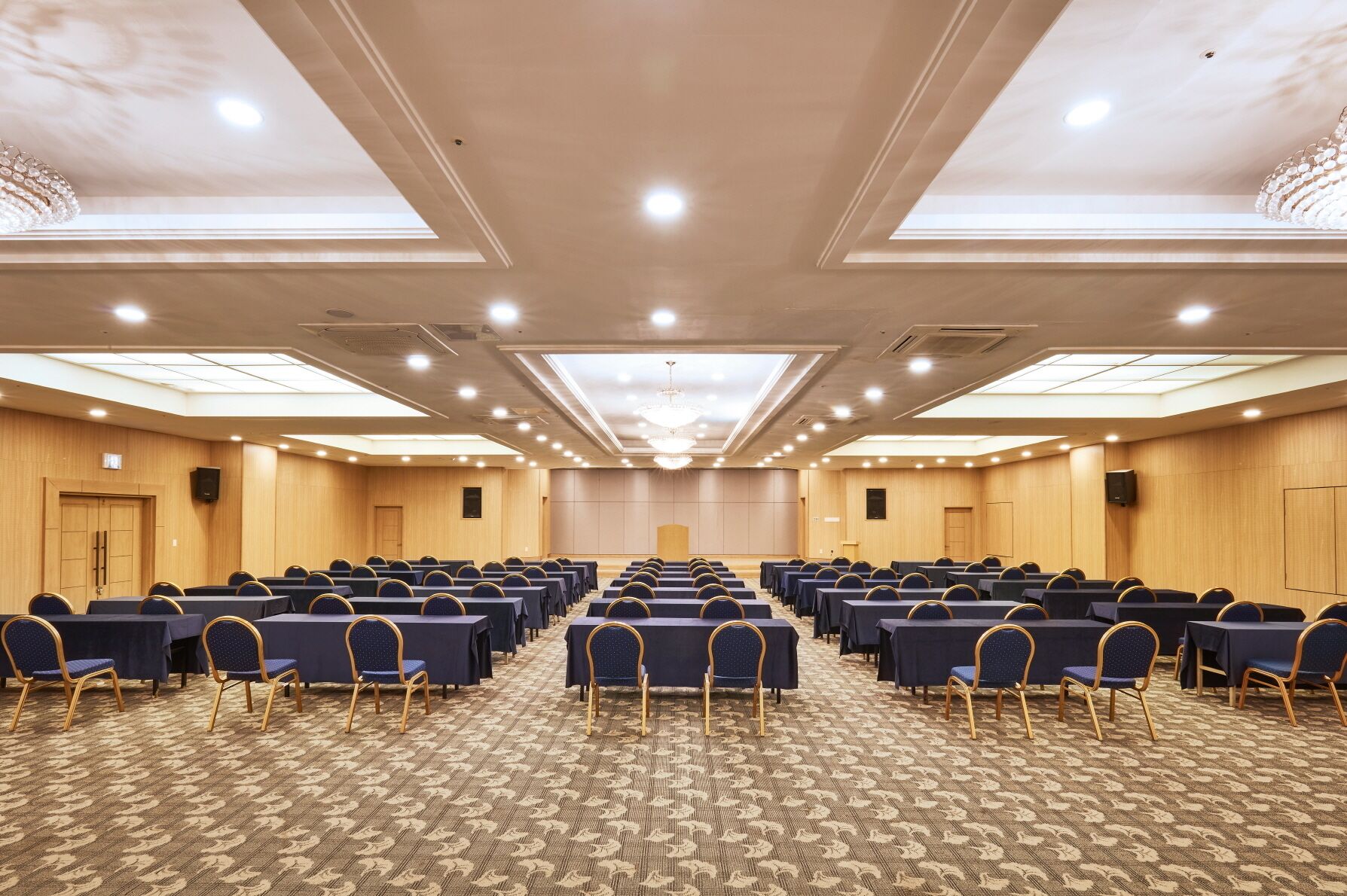 A conference room with rows of tables and chairs, a stage at the front, and a high ceiling with skylights.