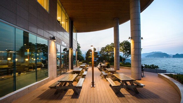 An outdoor dining area with wooden picnic tables and benches, a modern building with large windows, and a view of the water and hills in the background. - Kumho Tongyeong Marina Resort (Tongyeong)