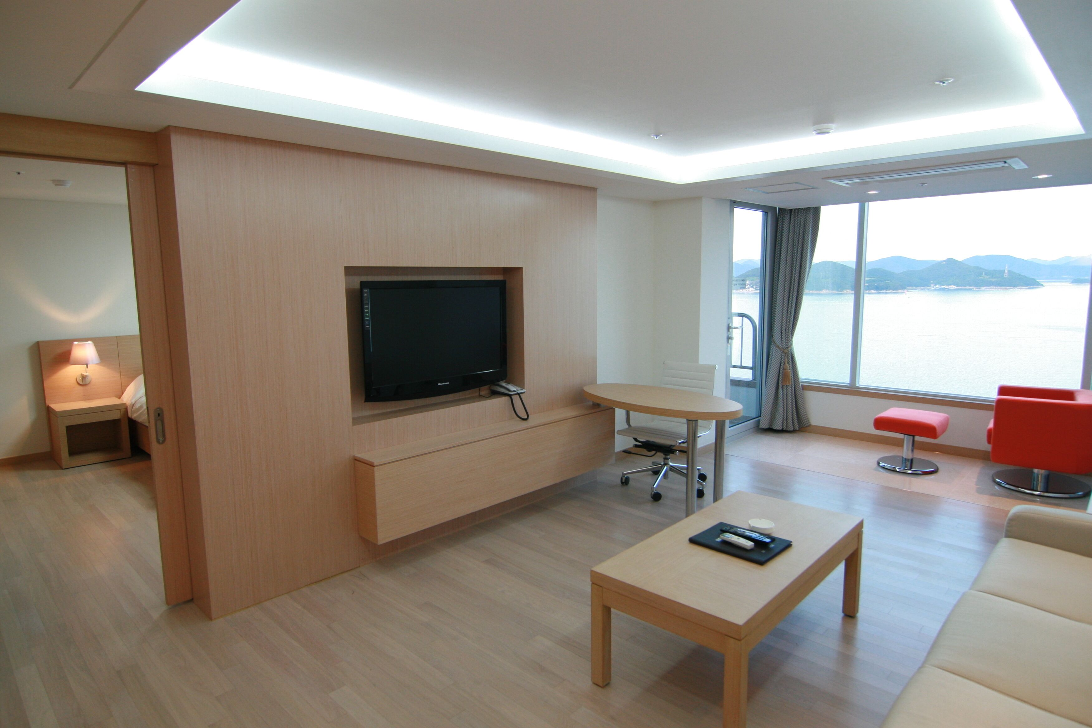 Una moderna camera d'albergo con una TV a schermo piatto fissata a un pannello di legno, un tavolino con un telecomando, una poltrona rossa e un'ampia finestra con vista su un corpo d'acqua.